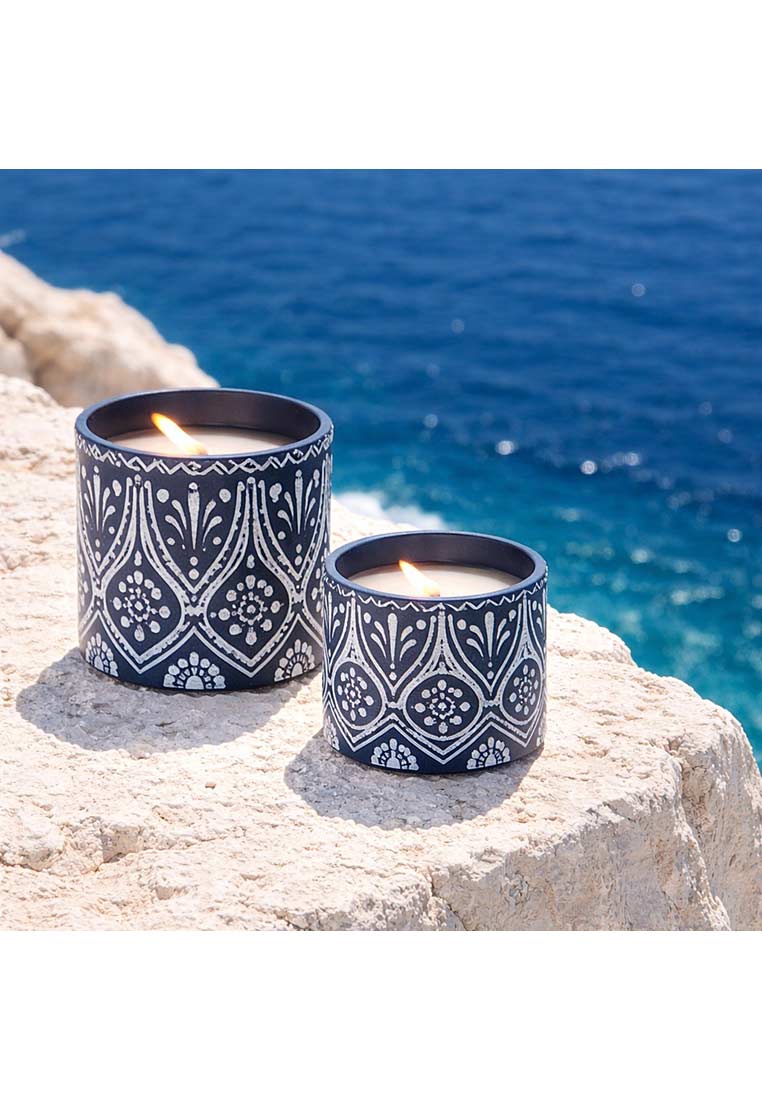 Zwei J-LINE Kerzen in blau und weiss mit Terracotta-Muster auf Stein mit Meer im Hintergrund, Frontansicht.
