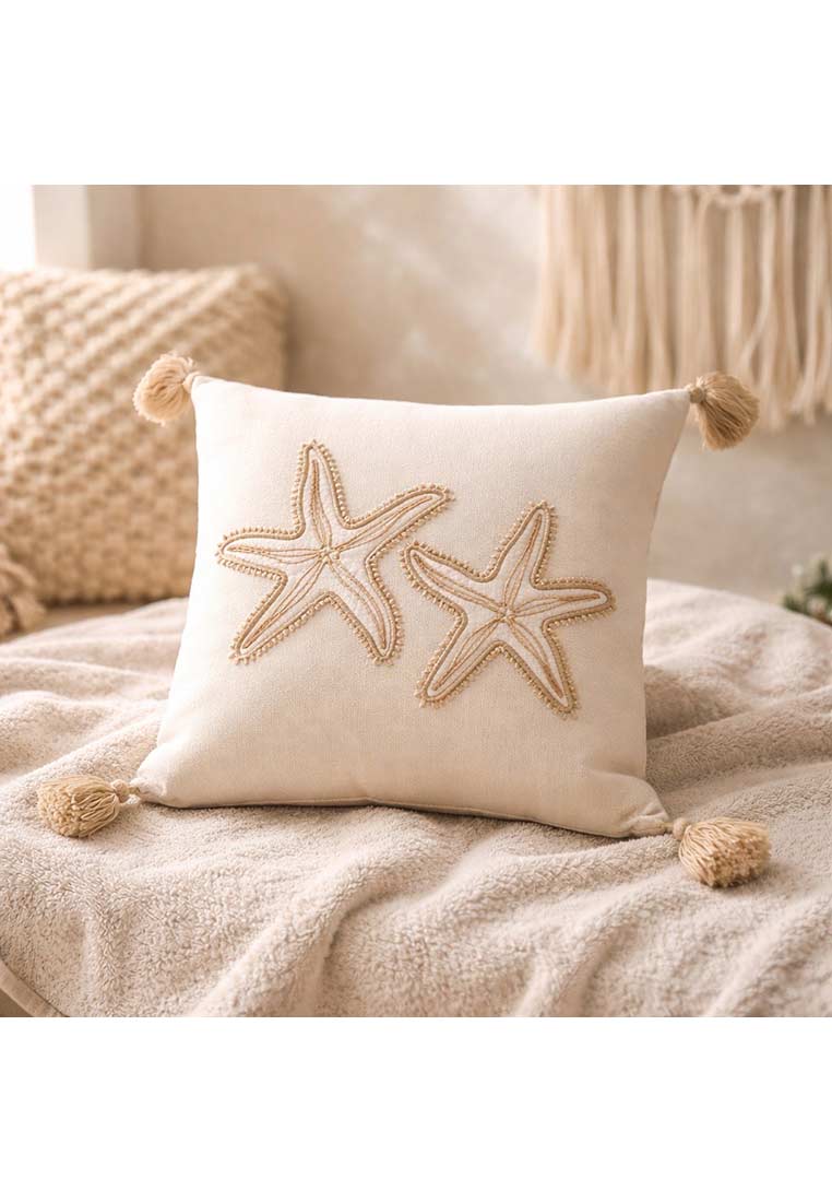 J-LINE Baumwollkissen Seestern in Beige mit zwei aufgestickten Seesternen und vier Quasten, Frontansicht. Das Kissen hat eine zarte Textur und ist auf einer weichen Decke platziert.