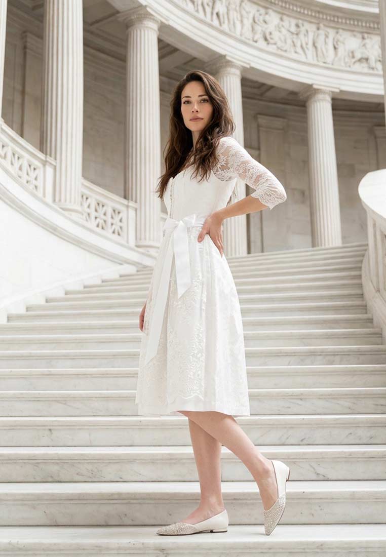 Stockerpoint Hochzeitsdirndl Josefina creme 70cm in einer eleganten Frontansicht auf einer Marmortreppe mit Spitzenärmeln und einer weißen Schleife.