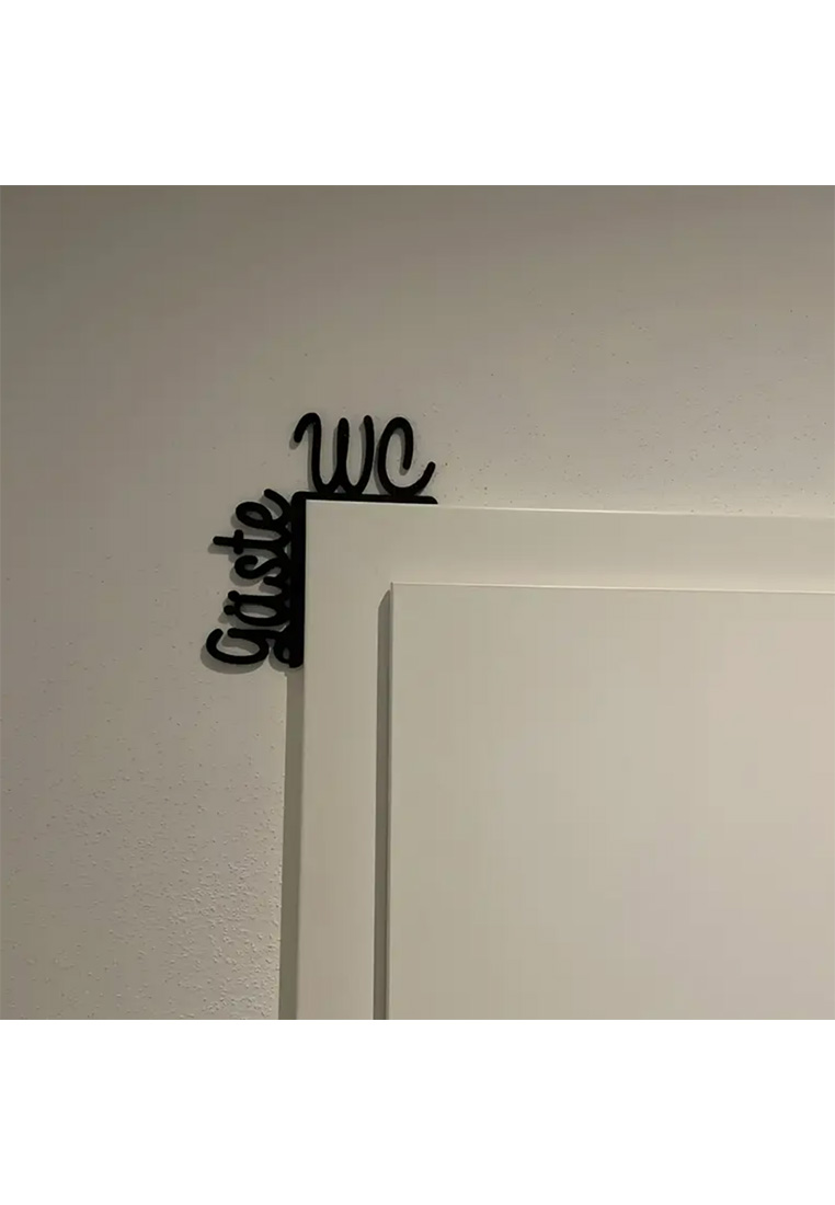 Eine schwarze Wanddekoration mit der Aufschrift "Gäste WC" in geschwungener Schrift, montiert an einer Wand in der Nähe einer Tür. Frontansicht.
