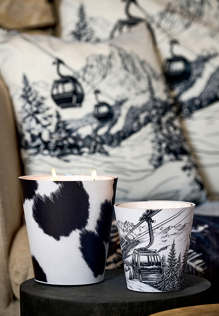 Pôdevache Chalet Chic Detailaufnahme mit Duftkerzen in Kuhfell- und Gondelbahn-Design, stimmungsvolles Wintermotiv vor passenden Kissen.