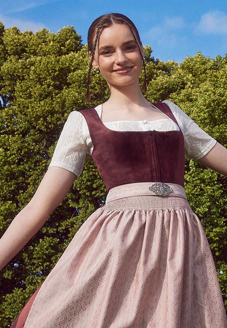 Frontansicht des Krüger Dirndl Aurela in Bordeaux mit einer 60cm langen Schürze und einem hellrosa Rock mit Muster, der mit einem breiten Band und einer Brosche verziert ist.