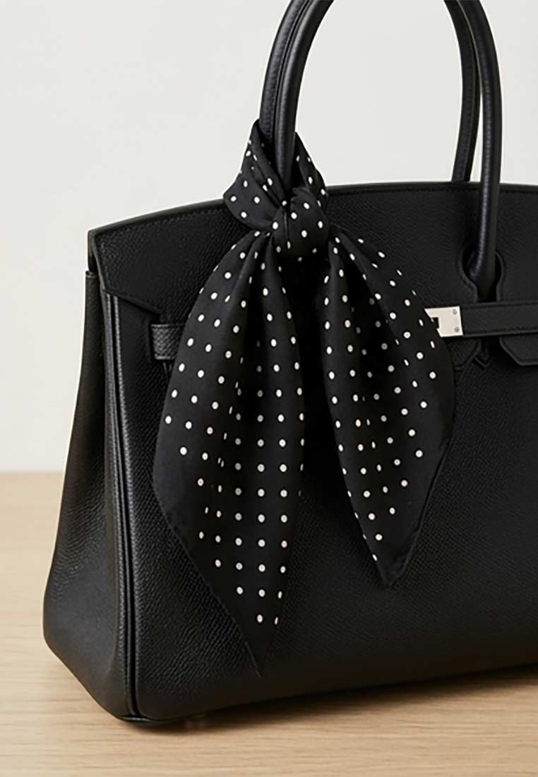 Schmuckzauber Nickituch Pünktchen schwarz aus Polyester, Frontansicht mit weißem Punktmuster und geknoteter Schleife um eine schwarze Handtasche.