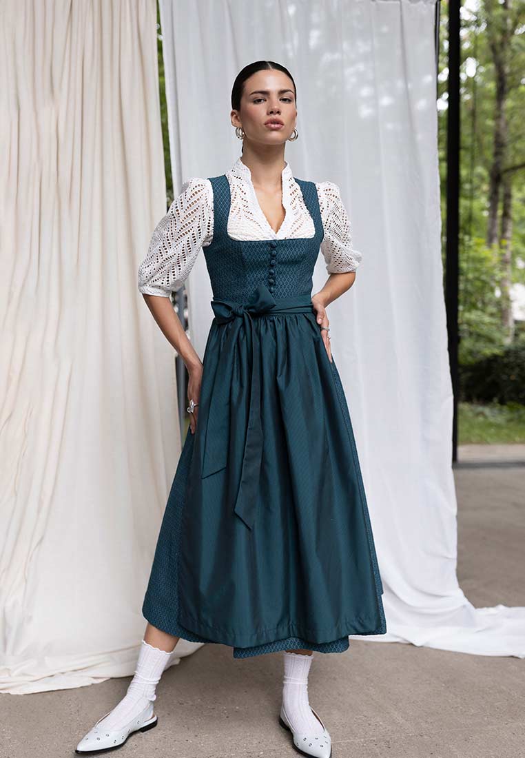 Madame Marie Dirndlbluse Norelin weiss, Frontansicht einer Frau in einem dunkelblauen Dirndl mit weißer Bluse und einer Schleife an der Taille. Das Dirndl hat einen durchgehenden Knopfverschluss.