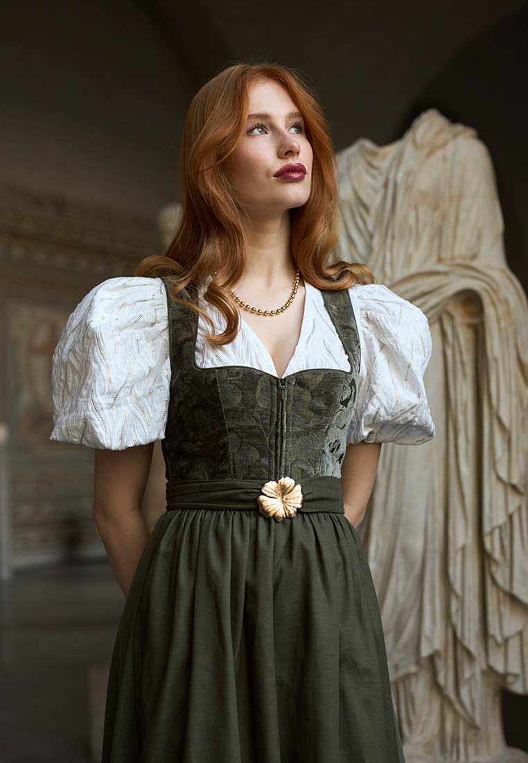 Madame Marie Dirndl Velirina Samt thymian 75 cm. Frontansicht eines Dirndls in Thymian-Grün mit weißer Bluse und Blumenmuster-Mieder. Das Dirndl hat einen Reißverschluss vorne und einen dekorativen Bund mit floraler Brosche.
