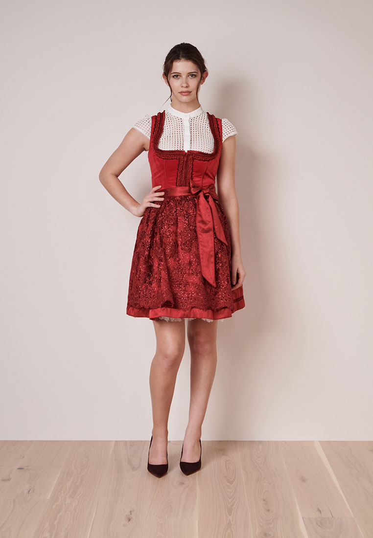 Krüger Dirndl Elma rot, eine Frau in vollständiger Frontansicht trägt dieses rote Dirndl mit einem Satinband in der Taille und einem Spitzenmuster auf dem Rock.